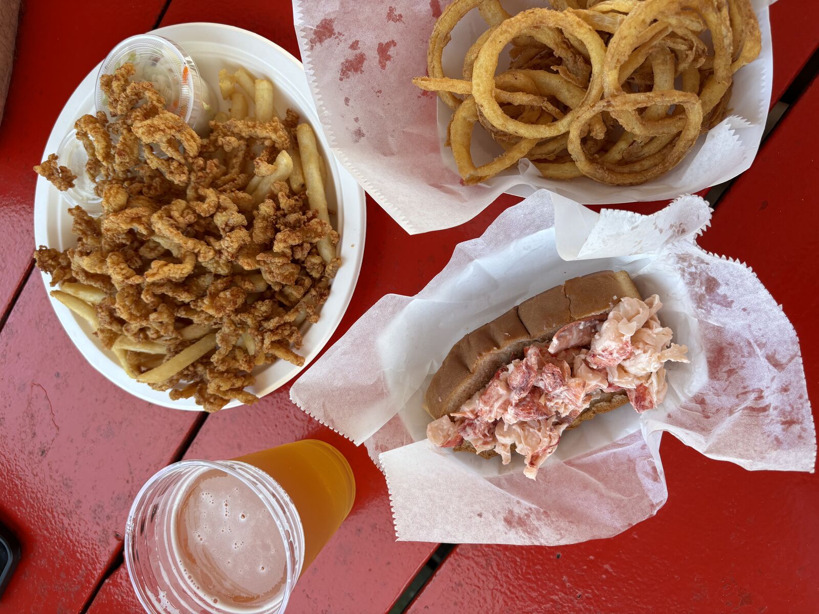 Lobster roll, fried clams with fries, onion rings, and a cold beer on a red picnic table at Cape Pier Chowder House